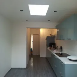 Nowoczesna kuchnia po remoncie: szare panele podłogowe imitujące drewno, białe ściany, błękitne szafki kuchenne z białym blatem, oświetlenie sufitowe punktowe i okno dachowe.