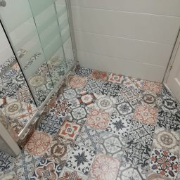 Mariusz Markowski - Wzorzyste płytki podłogowe w łazience, ułożone przy kabinie prysznicowej z częściowo matową szybą i białymi płytkami ściennymi oraz drzwiami.