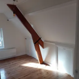 Poddasze po remoncie z drewnianą belką konstrukcyjną, panele podłogowe w odcieniach brązu, białe ściany i grzejnik pod oknem.