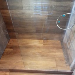 Mariusz Markowski - Kabina prysznicowa z drewnopodobnymi płytkami na podłodze i szarymi płytkami imitującymi marmur na ścianach, widoczny srebrny zestaw prysznicowy z niebieskimi akcentami.