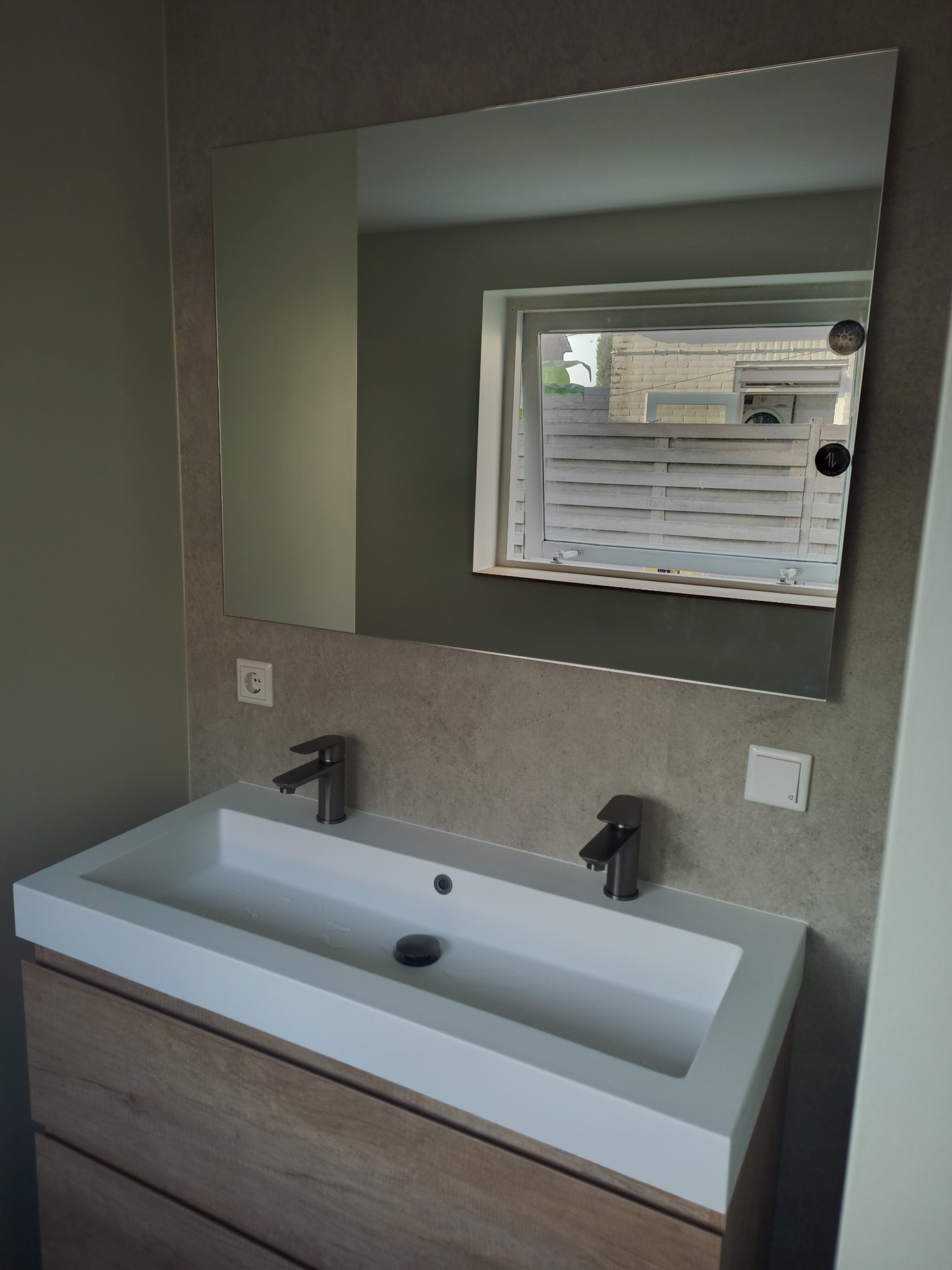 Nowoczesna umywalka nablatowa z dwiema bateriami w odcieniach szarości, z szafką z drewnopodobnym dekorem, nad nią lustro odbijające okno z widokiem na białe ogrodzenie i budynek.