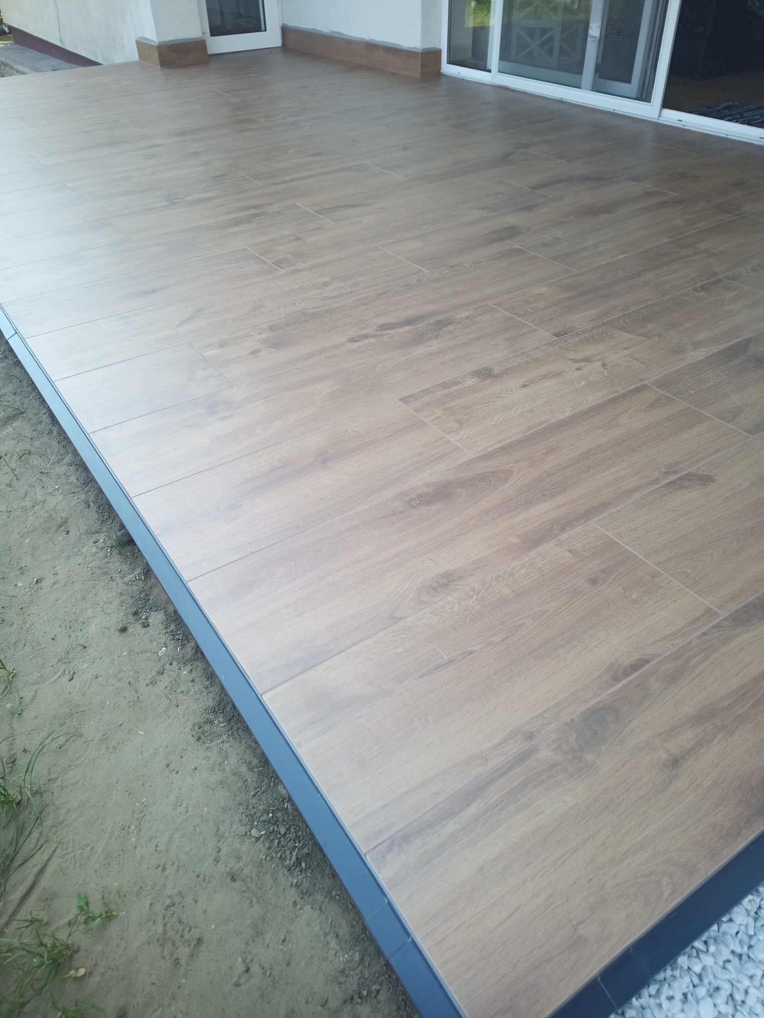 Ułożona podłoga z płytek imitujących drewno na zewnątrz budynku, widoczne szare obrzeże i fragment elewacji z oknami przesuwnymi.