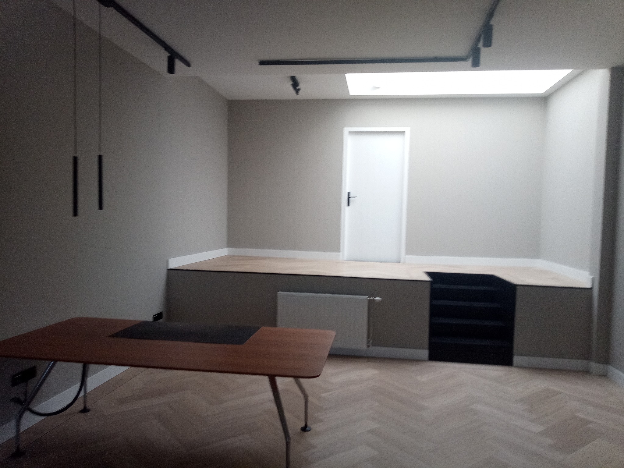 Wnętrze pokoju z podwyższoną podłogą wykończoną parkietem w jodełkę, białe drzwi, grzejnik i drewniany stół z czarnym blatem w lewym dolnym rogu.