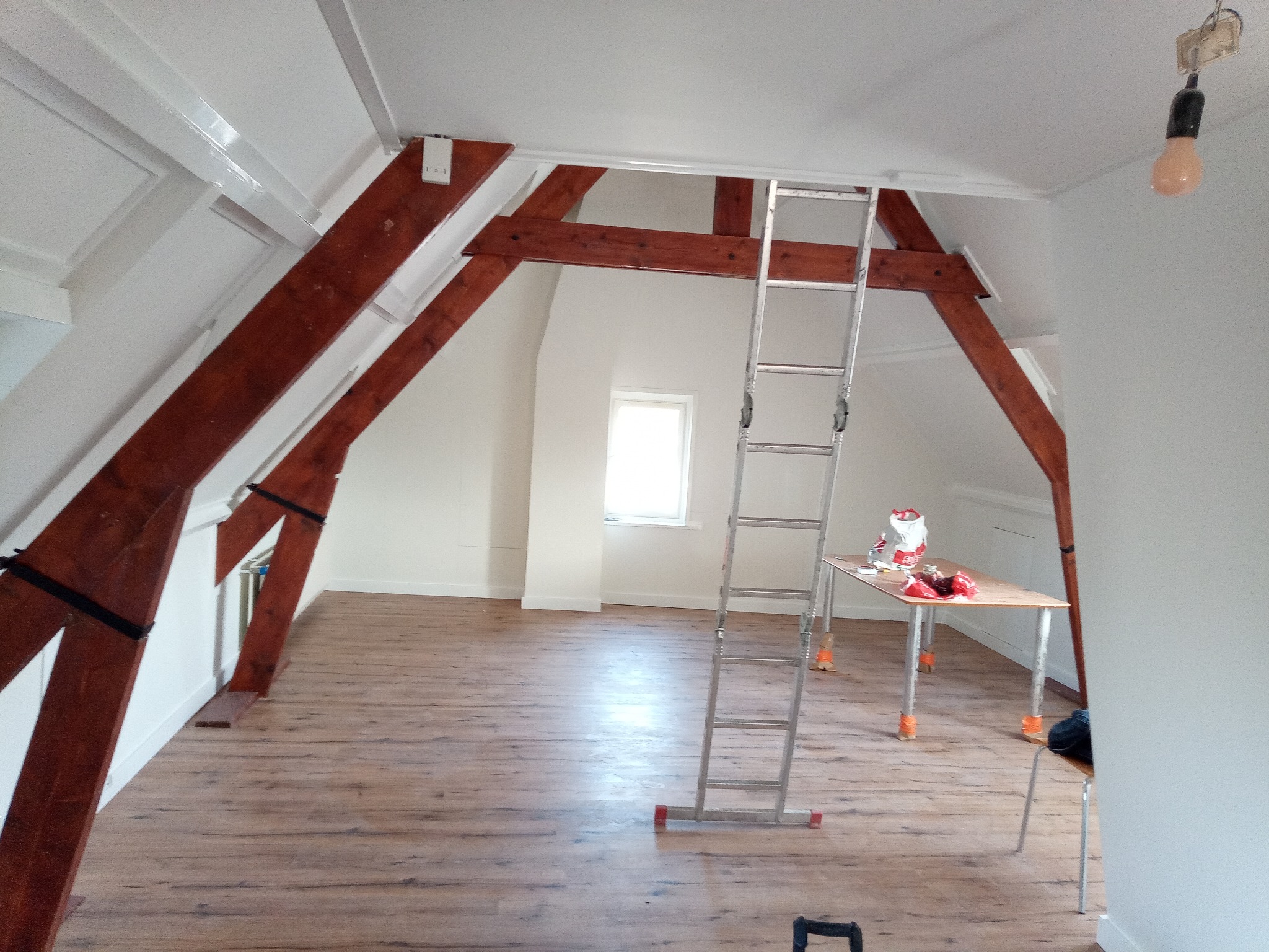Poddasze po malowaniu z drewnianymi belkami, aluminiowa drabina oparta o ścianę, stolik z farbami, surowe oświetlenie żarówką