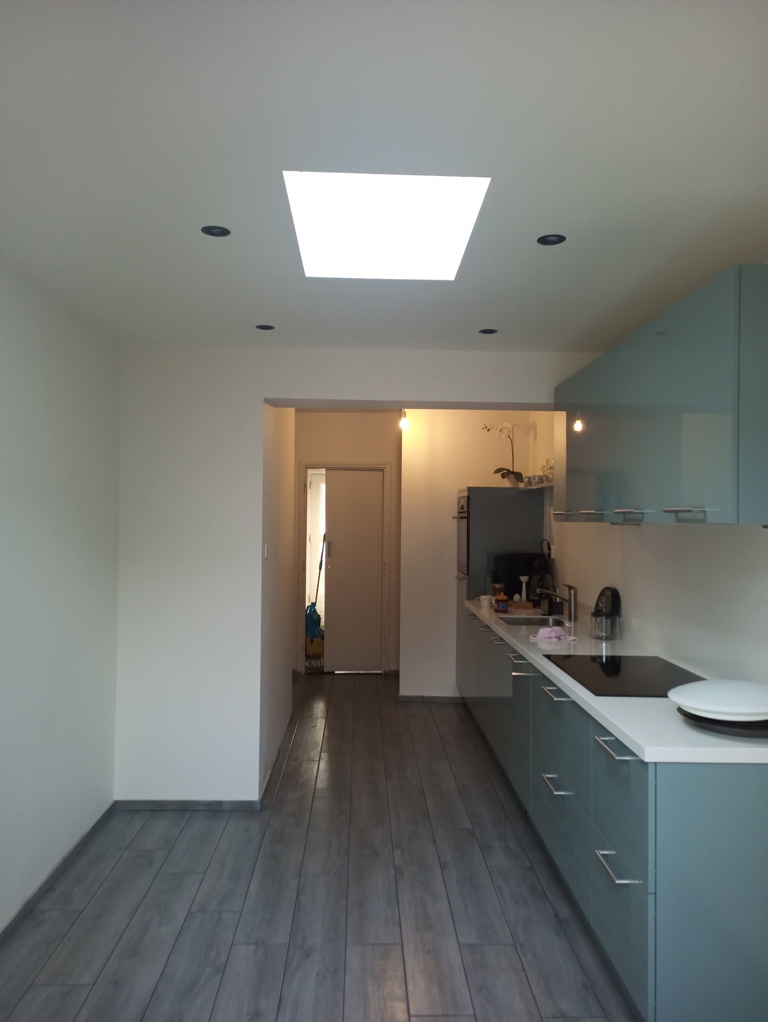 Nowoczesna kuchnia po remoncie: szare panele podłogowe imitujące drewno, białe ściany, błękitne szafki kuchenne z białym blatem, oświetlenie sufitowe punktowe i okno dachowe.