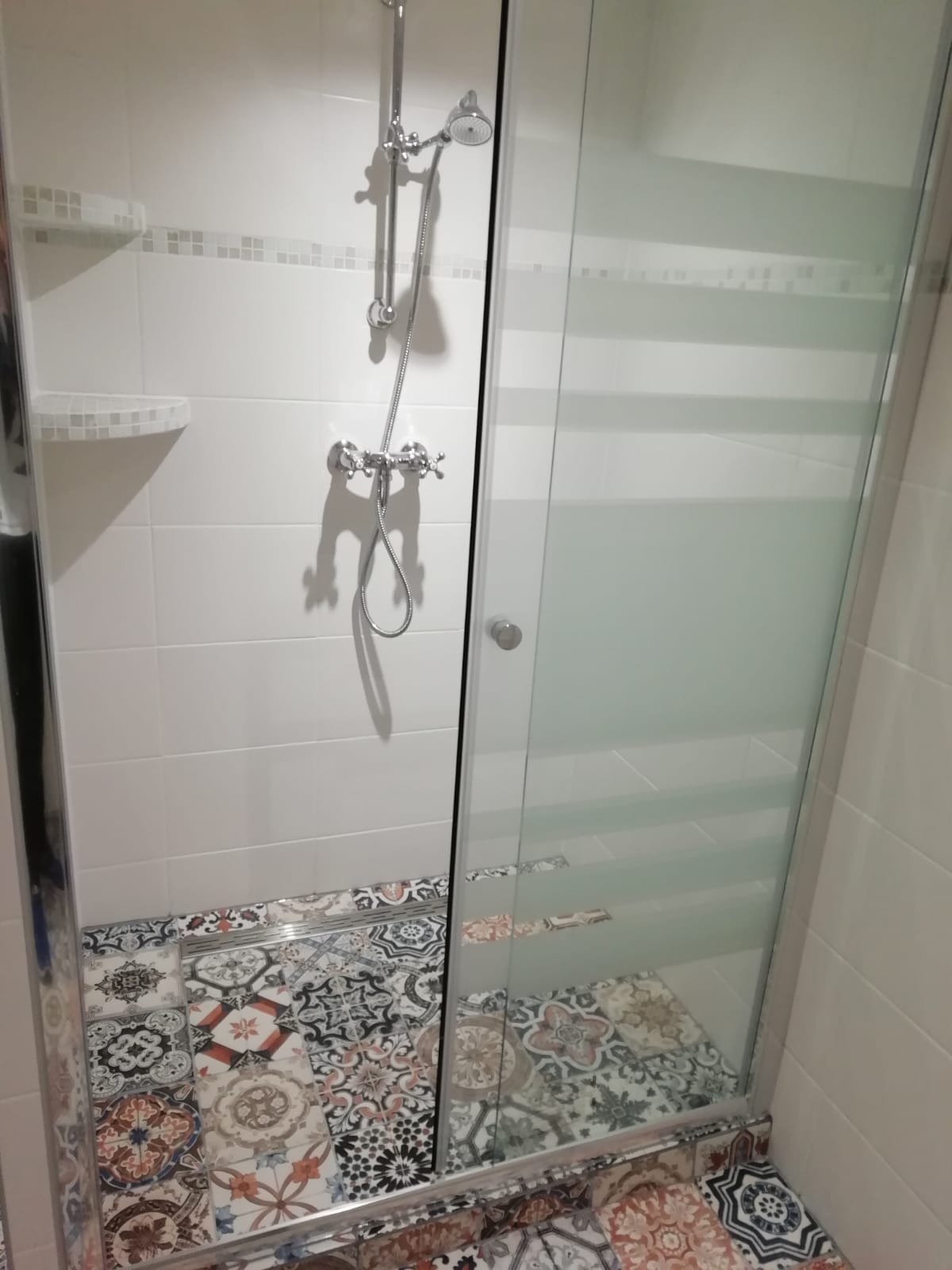 Kabina prysznicowa z chromowaną armaturą i wzorzystą podłogą z płytek ceramicznych, widoczne białe płytki na ścianach z dekoracyjnym pasem mozaiki.