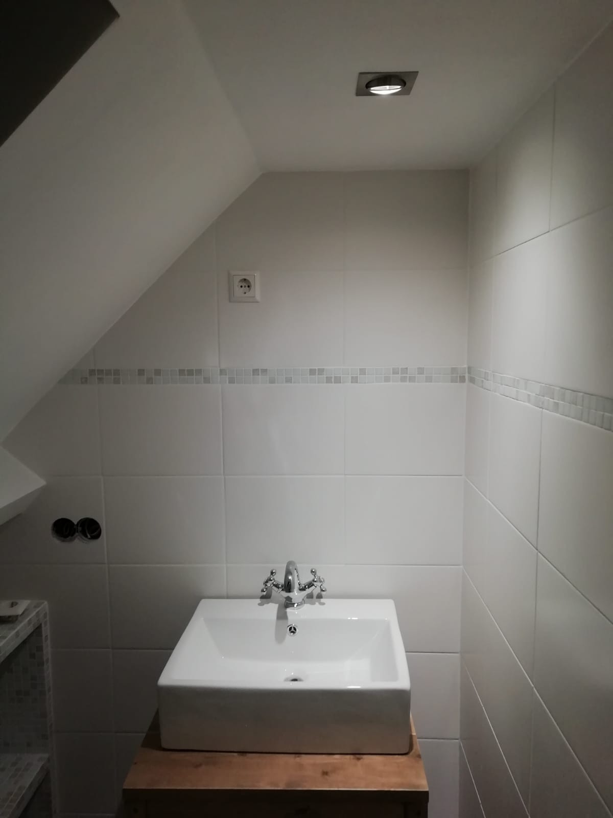 Biała, kwadratowa umywalka z chromowaną baterią w łazience na poddaszu, wykończonej białymi płytkami z dekoracyjnym pasem mozaiki i punktowym oświetleniem.