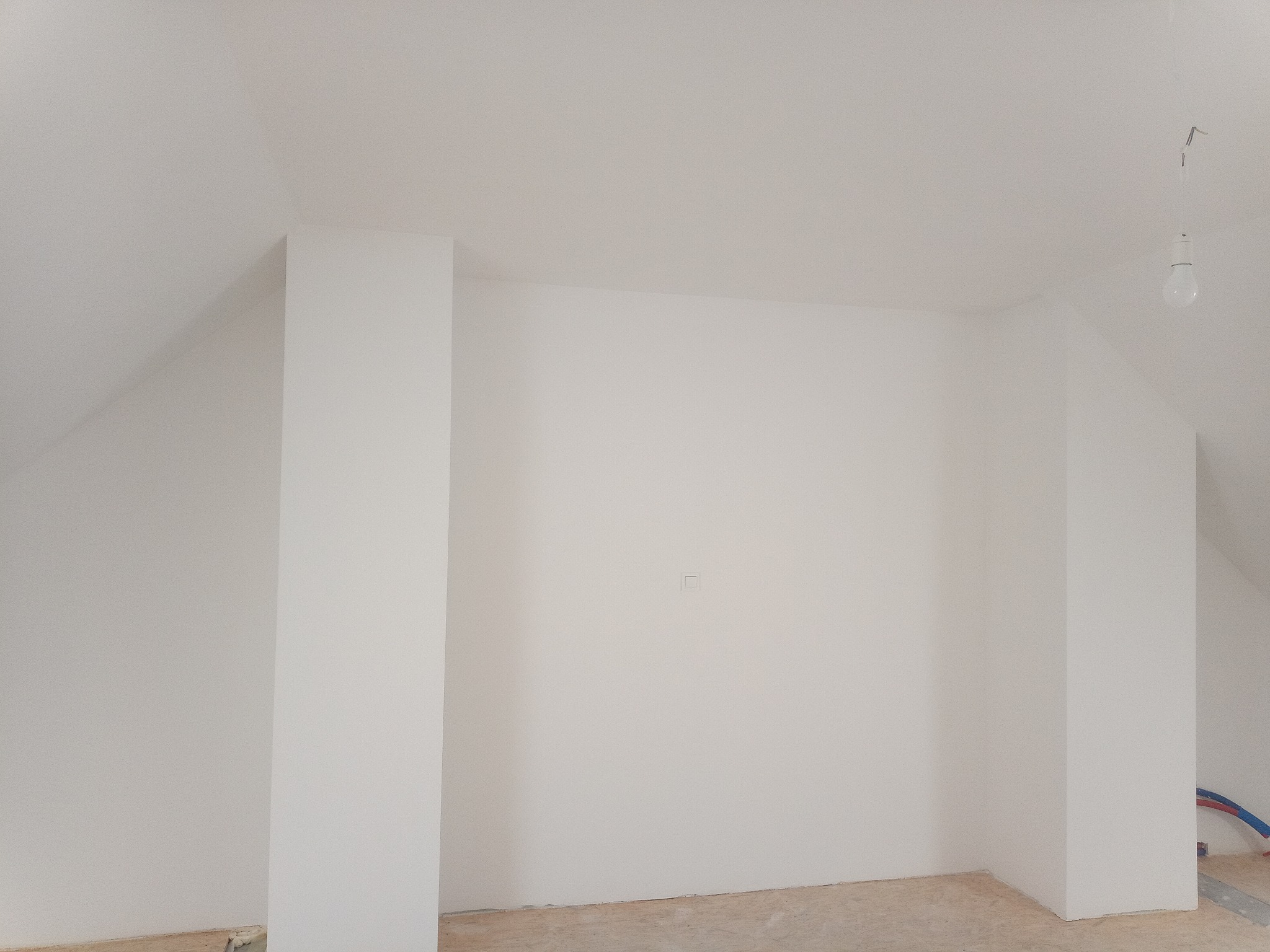 Poddasze po malowaniu na biało, widoczne słupy konstrukcyjne, gładkie ściany i sufit, na podłodze płyty OSB, widoczne fragmenty instalacji hydraulicznej.