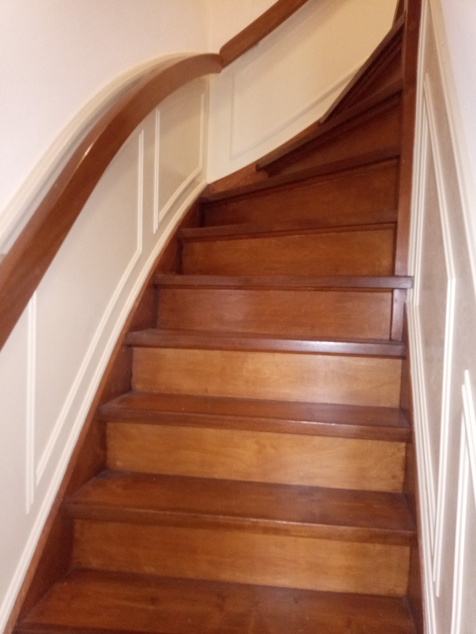 Drewniane schody z zakrętem i drewnianą balustradą, białe panele ścienne z dekoracyjnymi listwami.