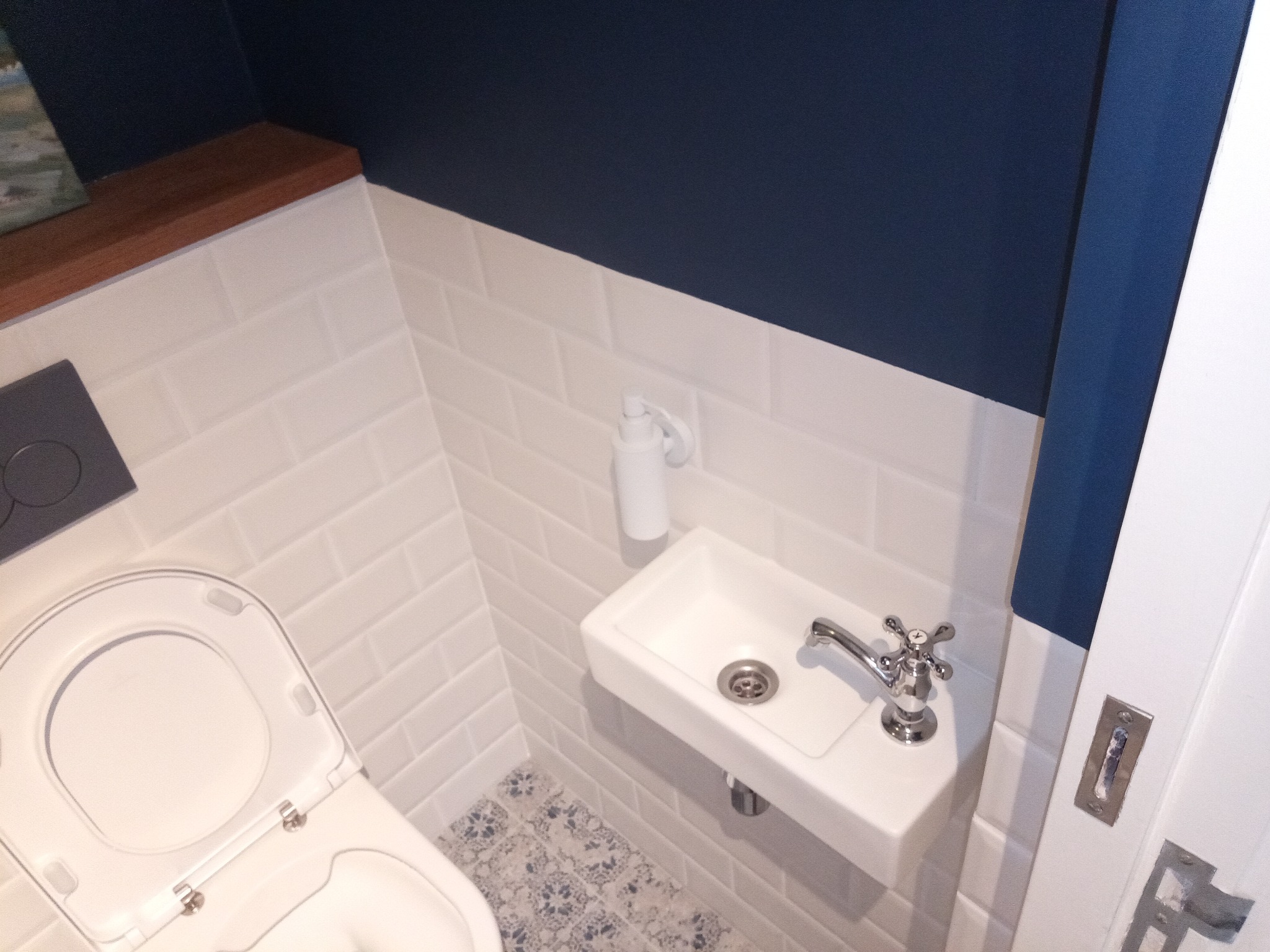 Nowoczesna, mała łazienka z białą armaturą, białymi płytkami ułożonymi w cegiełkę i granatową ścianą, akcentem drewnianym oraz wzorzystą podłogą.