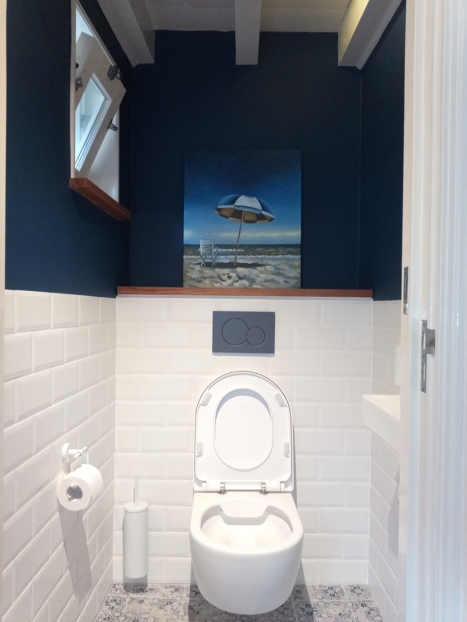 Nowoczesna toaleta z białymi płytkami, granatową ścianą, obrazem plaży i otwartym oknem.