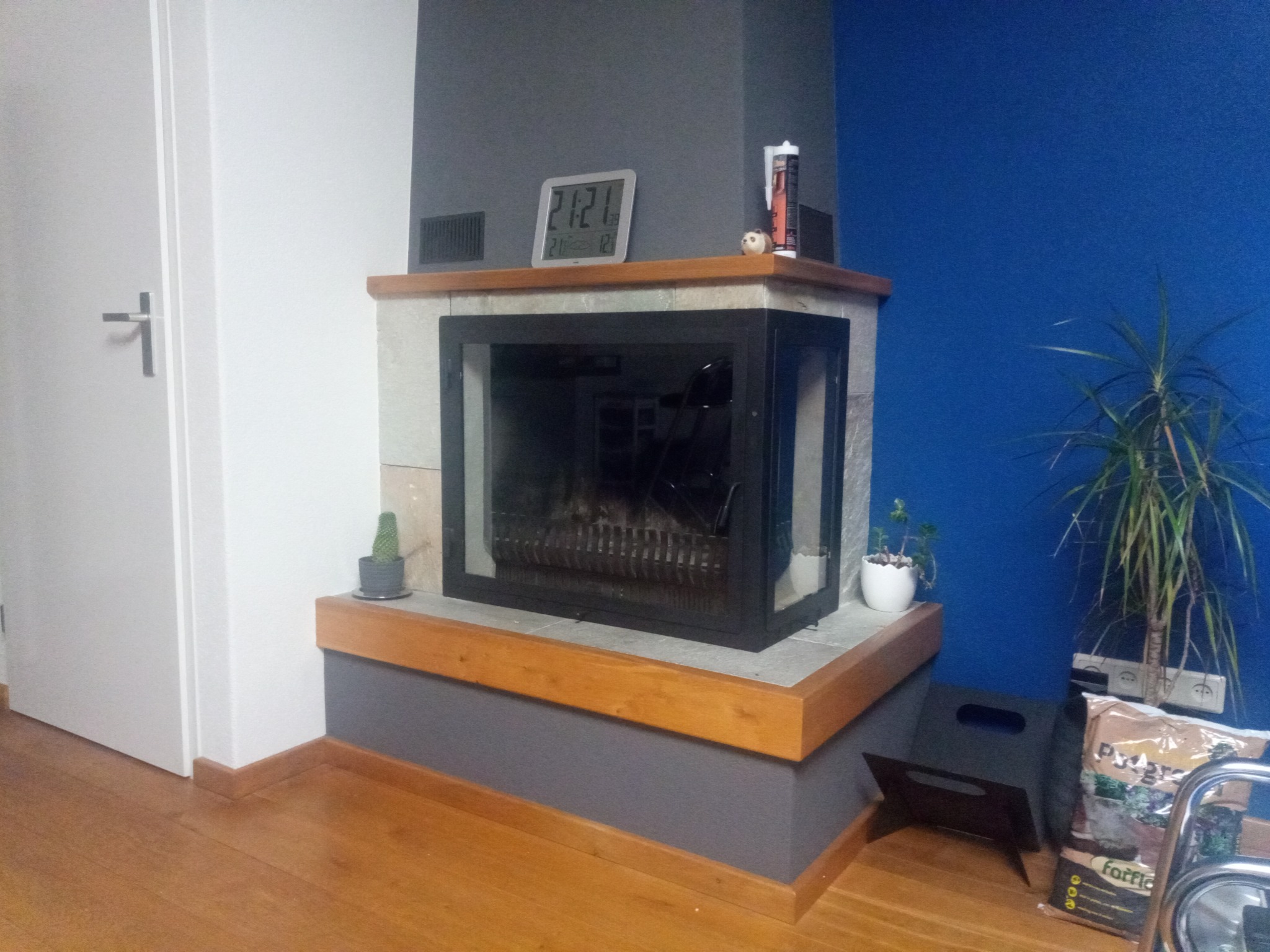 Nowoczesny kominek z kamienną obudową, drewnianymi elementami i czarną ramą, na którym stoi cyfrowy zegar i tuba silikonu, a obok, na tle niebieskiej ściany, roślina doniczkowa.