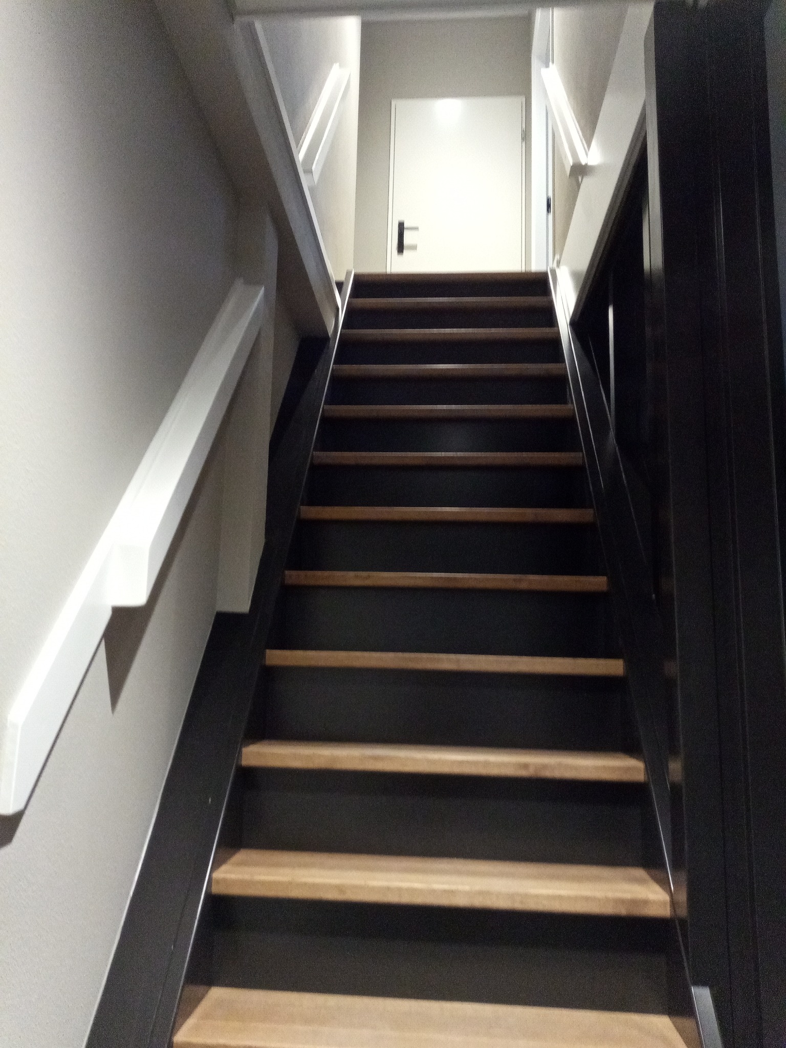 Klatka schodowa z drewnianymi stopniami i czarnymi podstopnicami, biała balustrada po lewej, czarna po prawej, białe drzwi w tle.