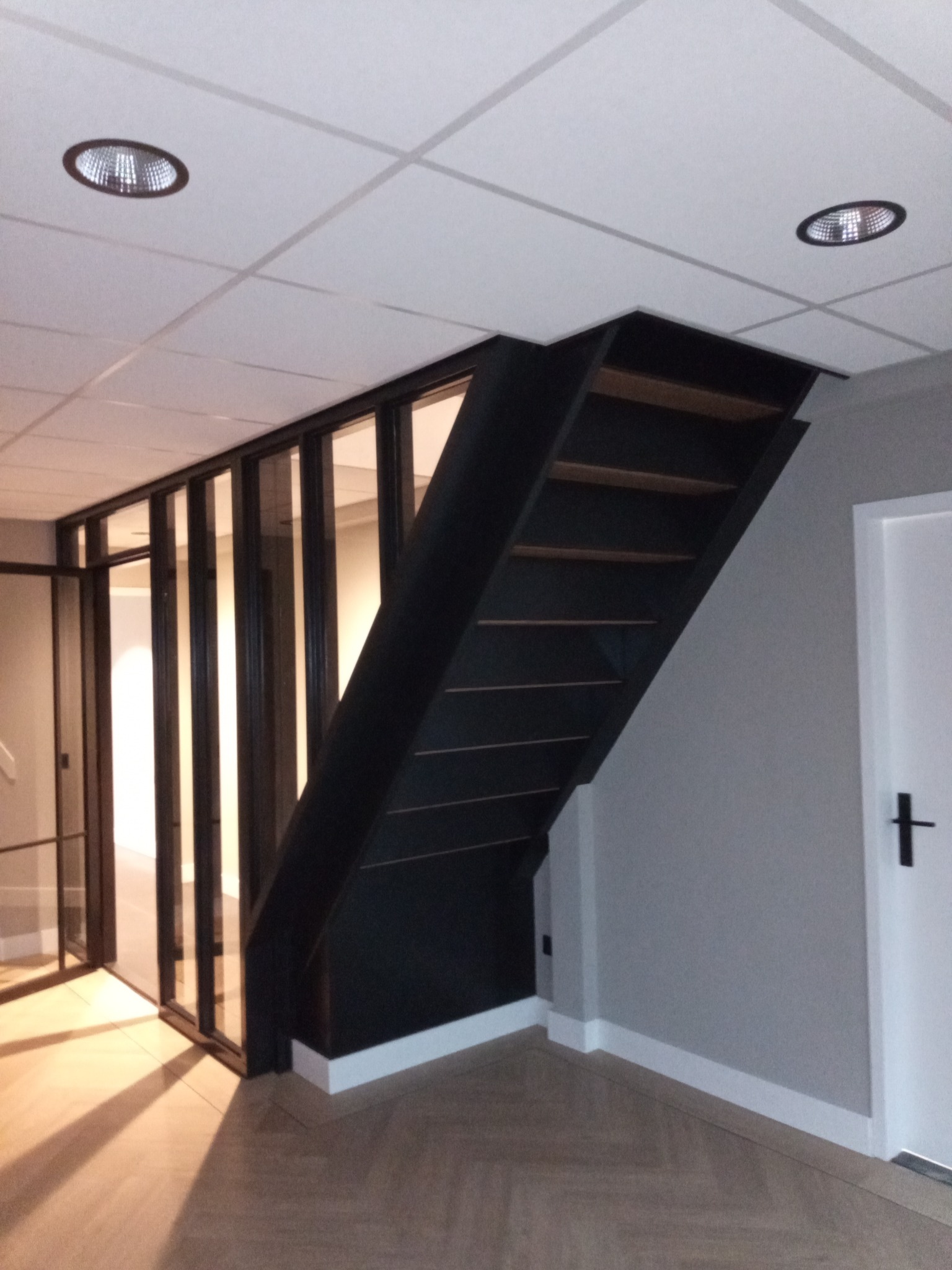 Nowoczesne schody w ciemnej kolorystyce z drewnianymi stopniami, szklana ścianka działowa, białe listwy przypodłogowe i parkiet w jodełkę.