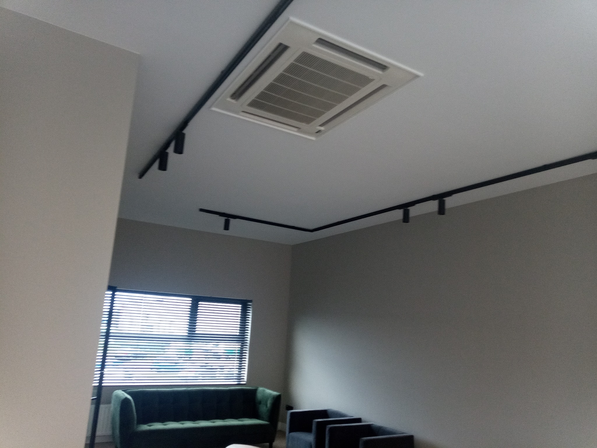 Klimatyzator kasetonowy w suficie podwieszanym z czarnymi szynami oświetleniowymi, widok na zieloną sofę i okno z żaluzjami w szarym pomieszczeniu.