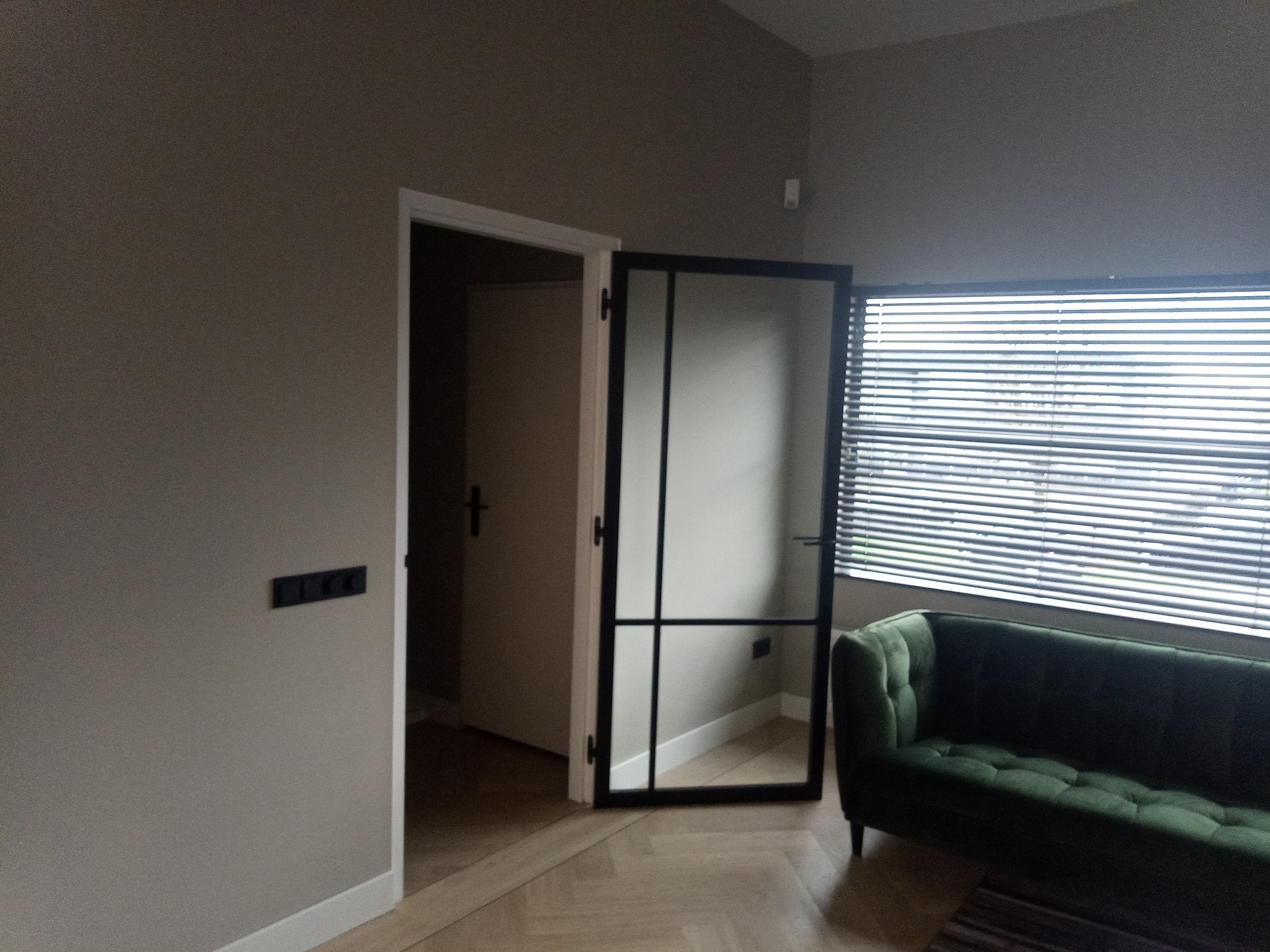 Wnętrze pokoju z szarą ścianą, otwartymi białymi drzwiami z czarną ramą, zieloną kanapą i drewnianą podłogą ułożoną w jodełkę.