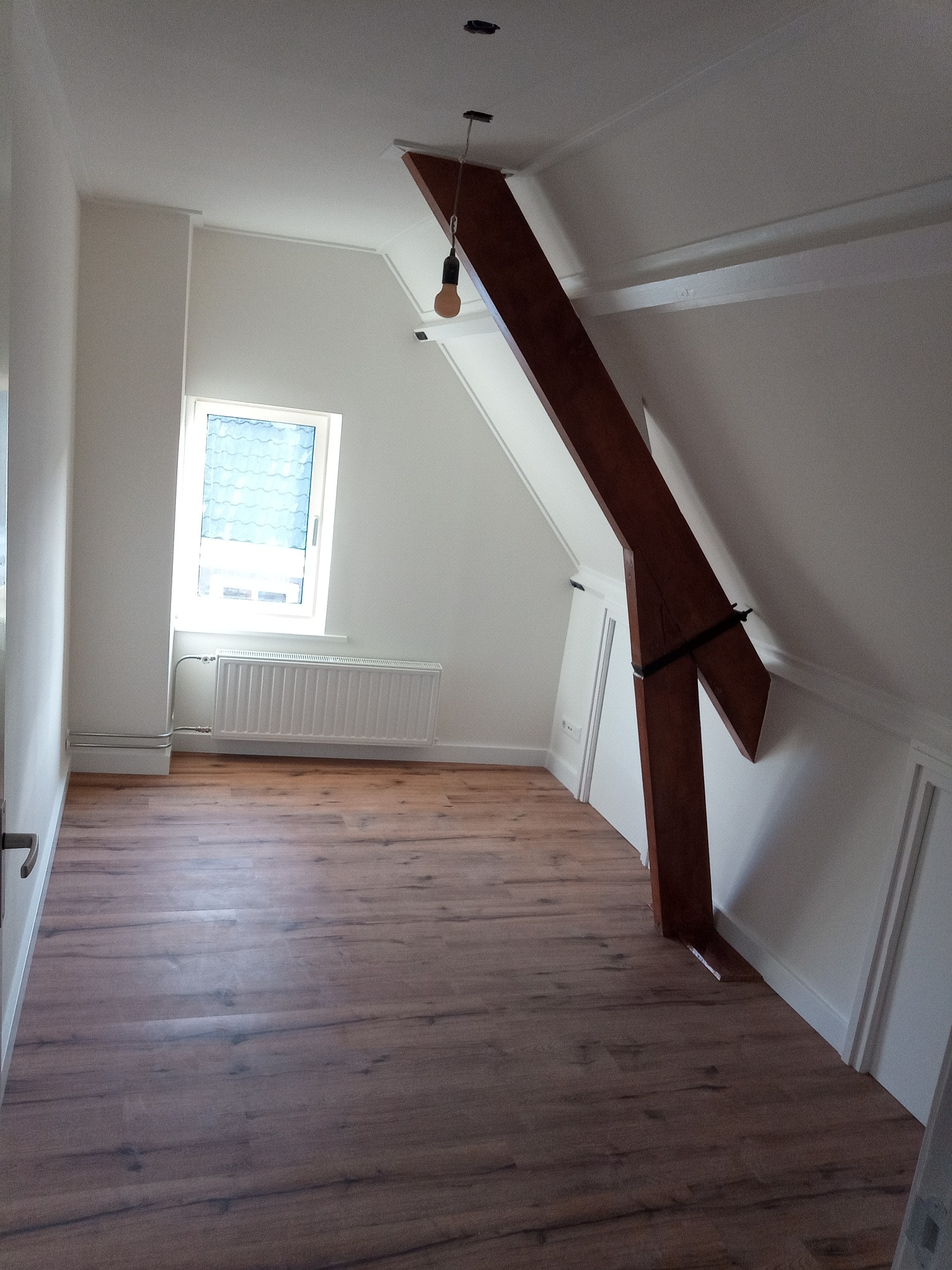 Poddasze po remoncie z drewnianą belką konstrukcyjną, świeżo ułożone panele podłogowe i pomalowane ściany na biało, widoczny grzejnik pod oknem.