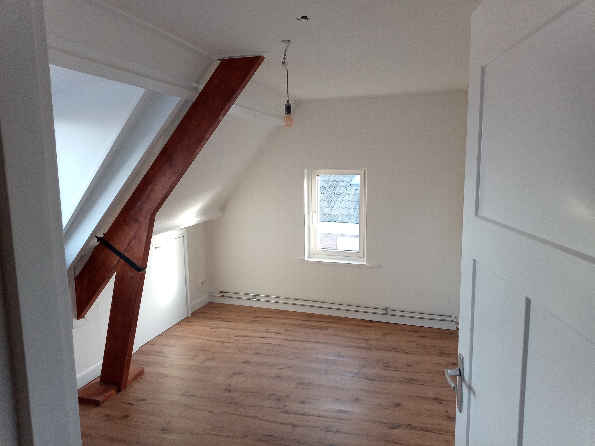 Poddasze po remoncie z drewnianą belką konstrukcyjną, panelami podłogowymi w odcieniach brązu i białymi ścianami oraz oknem dachowym.