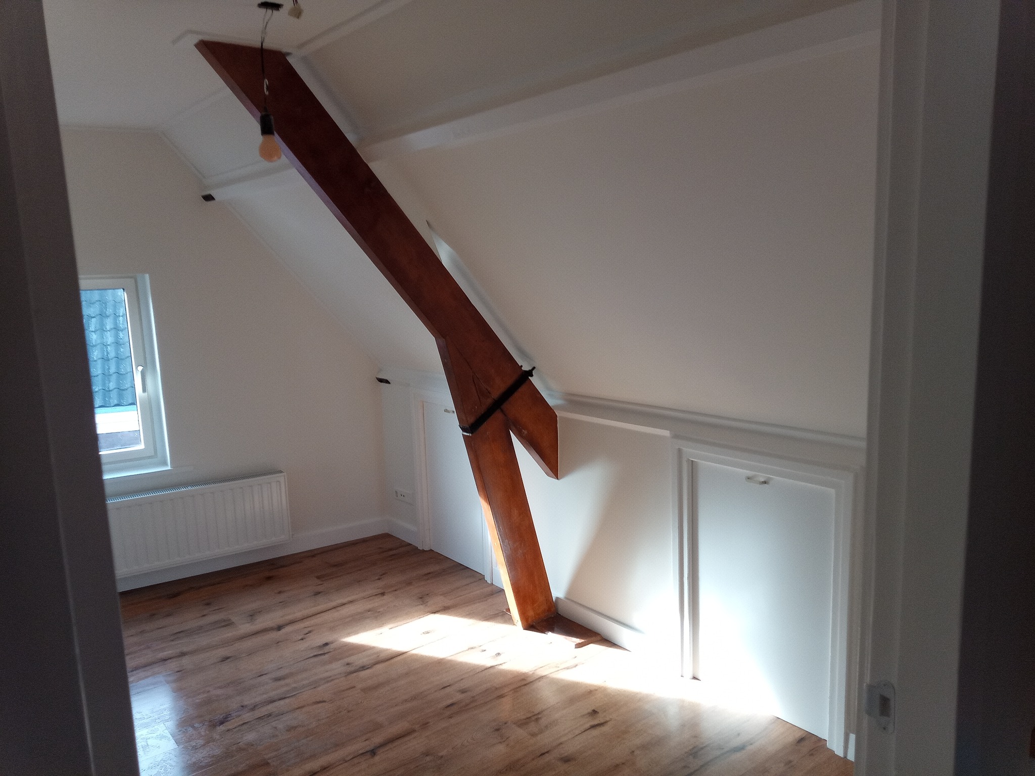 Poddasze po remoncie z drewnianą belką konstrukcyjną, panele podłogowe w odcieniach brązu, białe ściany i grzejnik pod oknem.