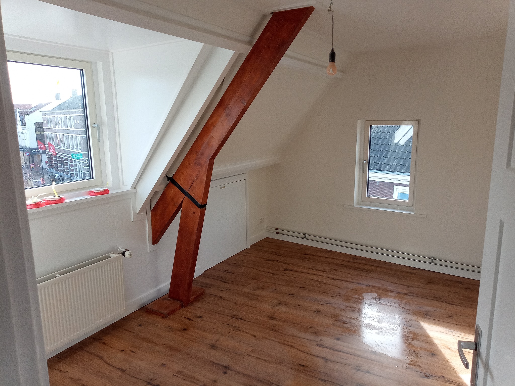Poddasze po remoncie w Chotomowie z nową podłogą z paneli, pomalowanymi na biało ścianami i drewnianą belką konstrukcyjną, widok na ulicę przez okno.