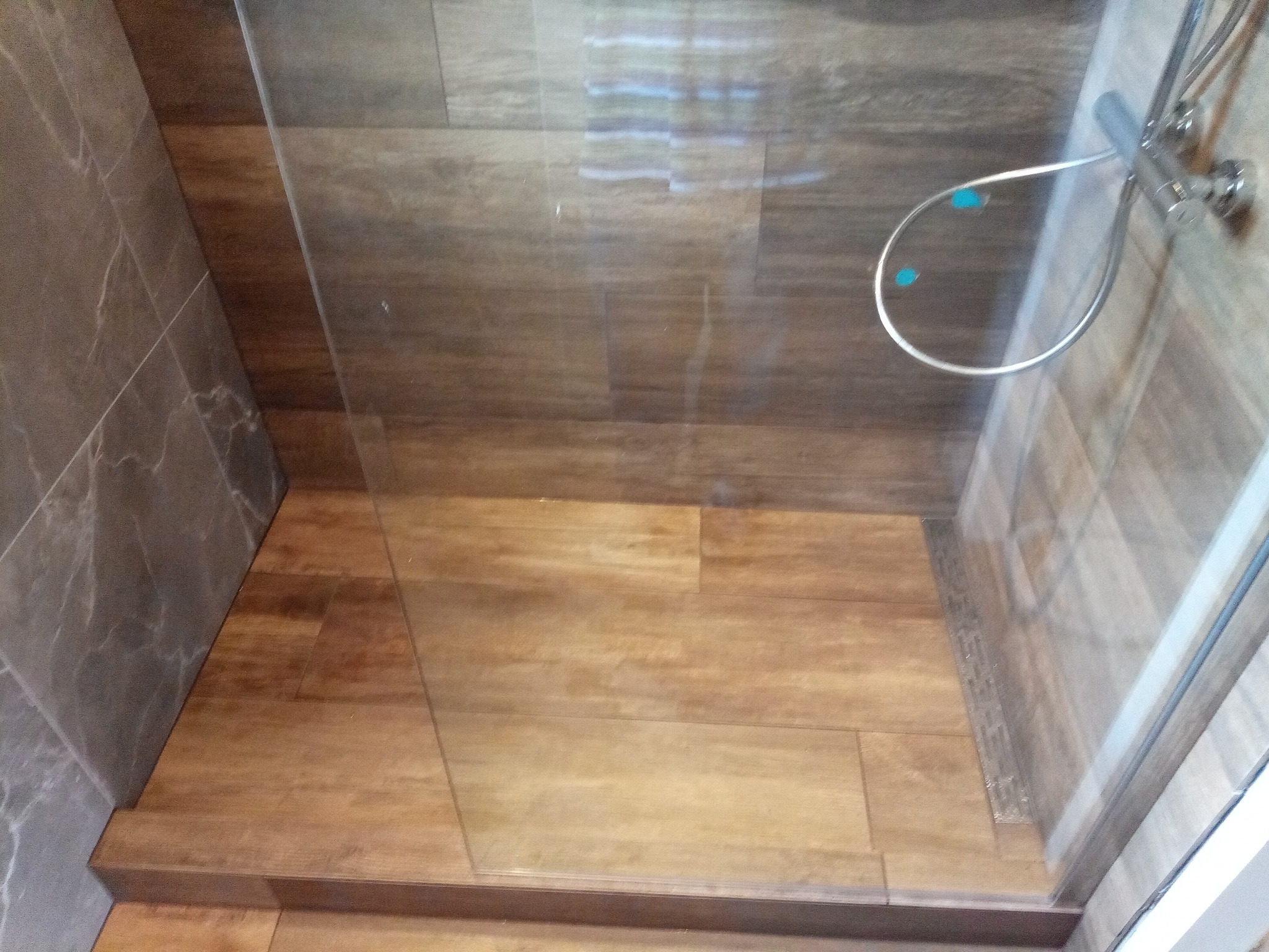 Kabina prysznicowa z drewnopodobnymi płytkami na podłodze i szarymi płytkami imitującymi marmur na ścianach, widoczny srebrny zestaw prysznicowy z niebieskimi akcentami.