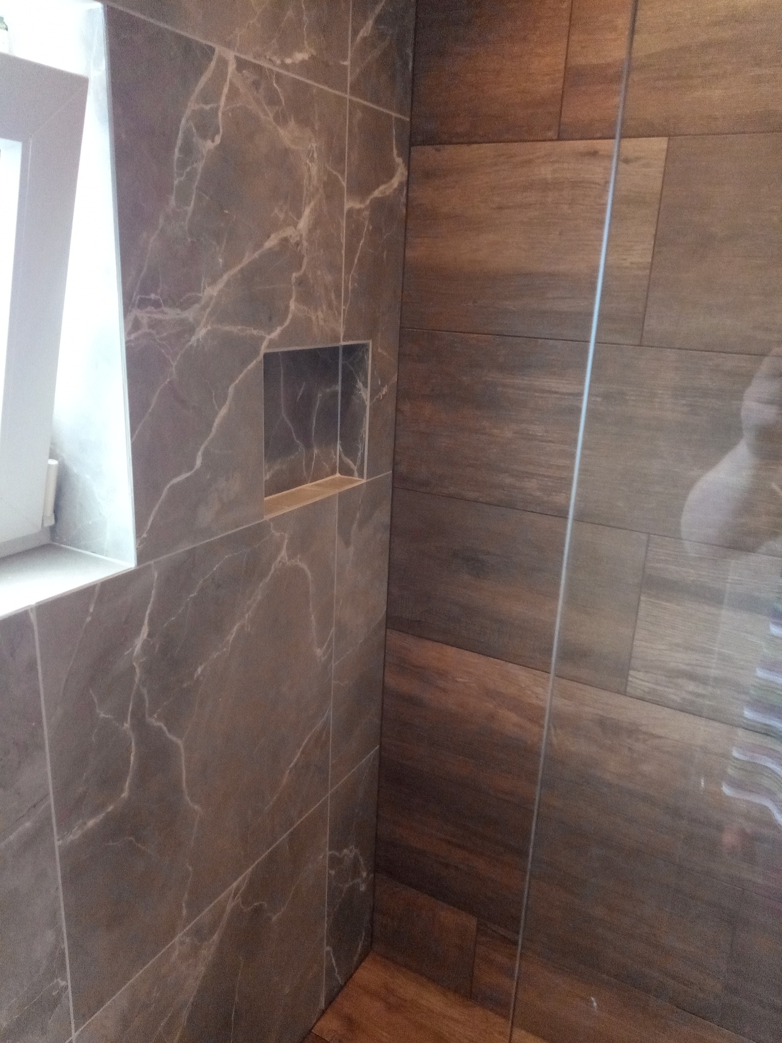 Ściana kabiny prysznicowej wyłożona brązowymi płytkami imitującymi drewno, sąsiadująca ze ścianą z szarych płytek imitujących marmur z wnęką.