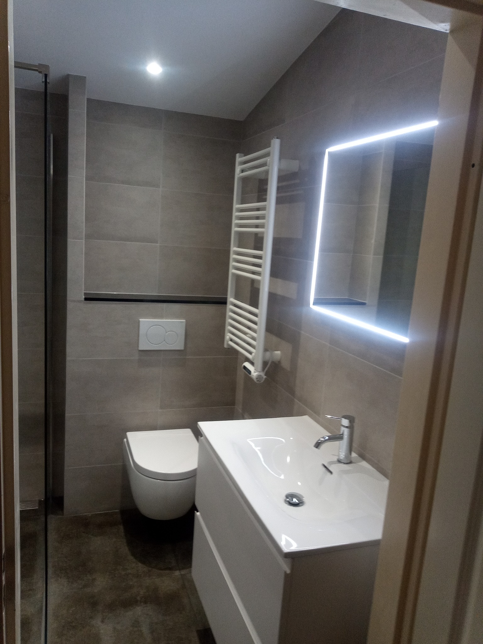 Nowoczesna łazienka w szarościach z podświetlanym lustrem, grzejnikiem drabinkowym i wiszącą toaletą.