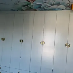 Biała szafa wnękowa z dekoracyjnymi, okrągłymi uchwytami w kolorze złotym, pod sufitem ozdobionym malowidłami kwiatowymi, oświetlona złotą lampą sufitową i wiszącą lampą z białymi kulami.