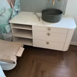 Łazienka z umywalką nablatową w kolorze grafitowym, armaturą w odcieniu złota i szafką z marmurowym blatem oraz ryflowanymi frontami szuflad, widoczny karton po ceramice i parkiet w jodełkę.