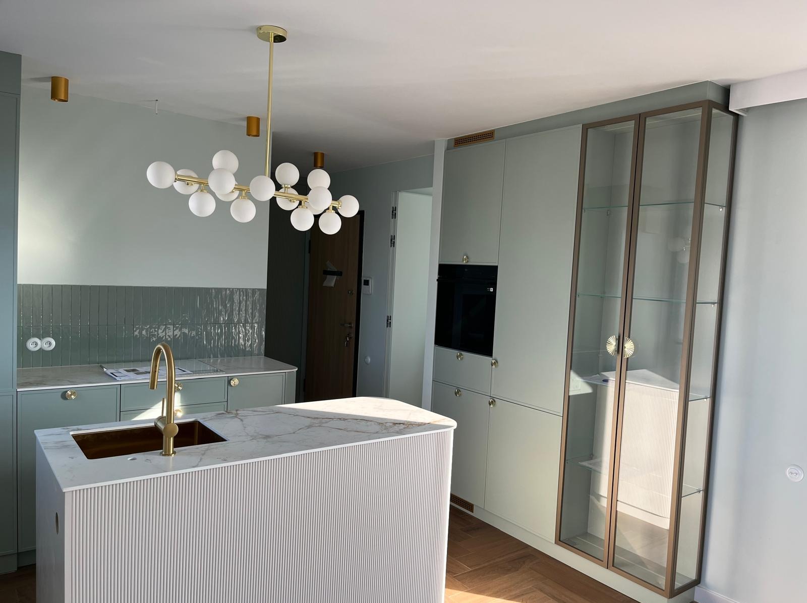 Nowoczesna kuchnia w odcieniach szarości i bieli z marmurowym blatem, złotą armaturą i designerskim oświetleniem, widok na wyspę kuchenną z ryflowanym frontem i zabudowę z witryną.