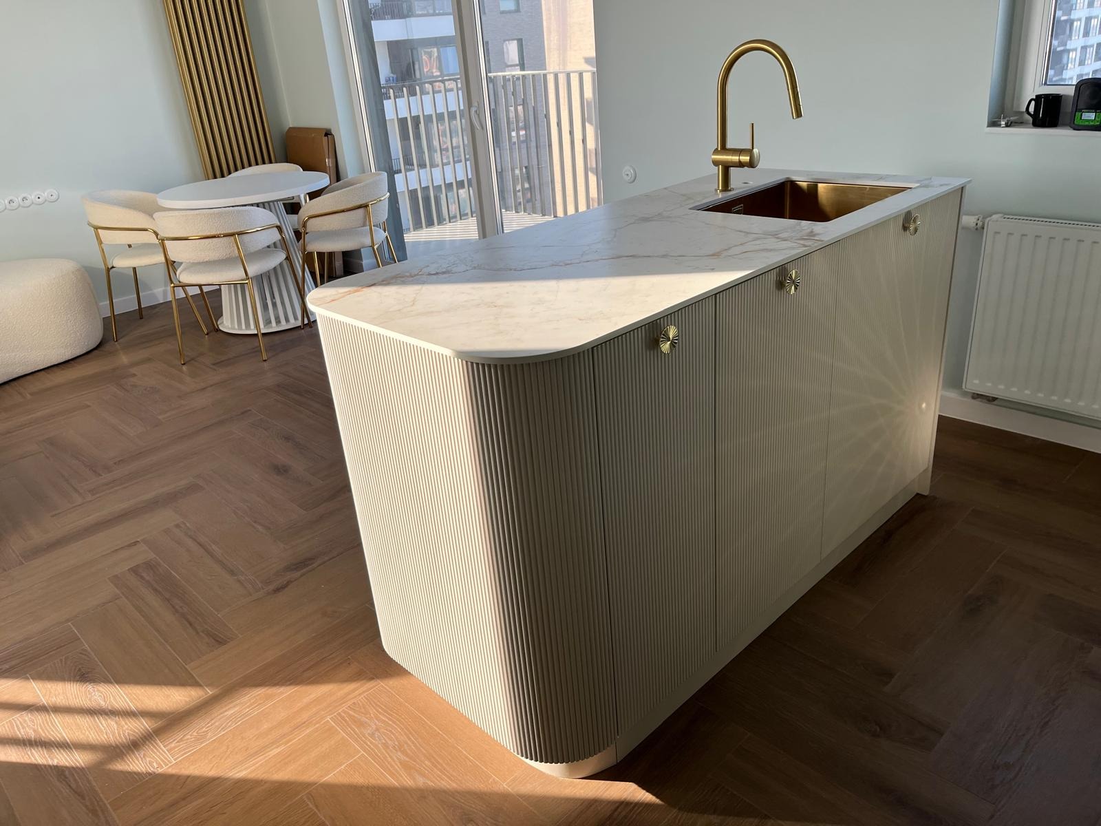 Wyspa kuchenna z marmurowym blatem, złota bateria i zlew, ryflowane fronty szafek, parkiet w jodełkę, jasne wnętrze z balkonem w tle.