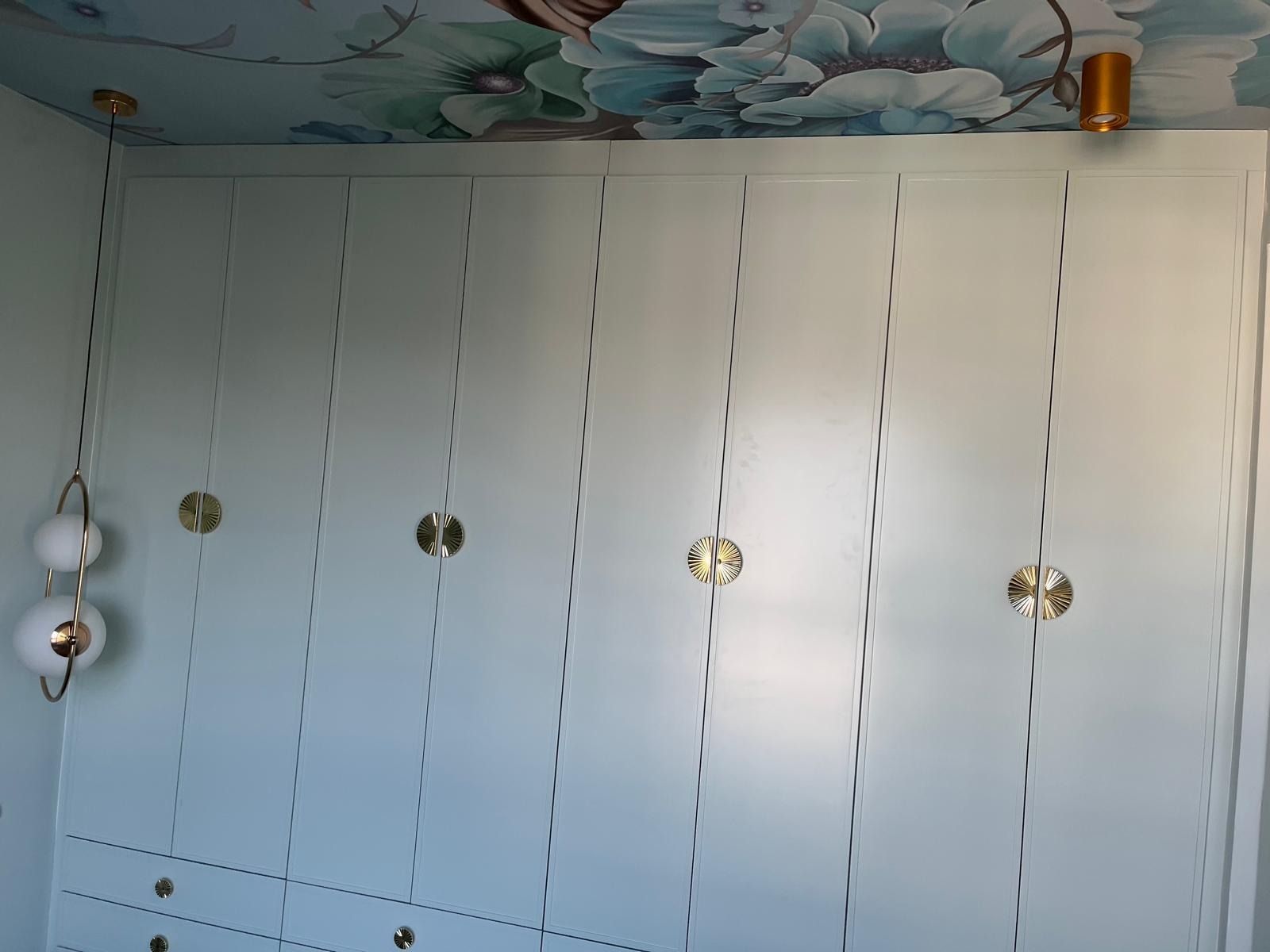 Biała szafa wnękowa z dekoracyjnymi, okrągłymi uchwytami w kolorze złotym, pod sufitem ozdobionym malowidłami kwiatowymi, oświetlona złotą lampą sufitową i wiszącą lampą z białymi kulami.