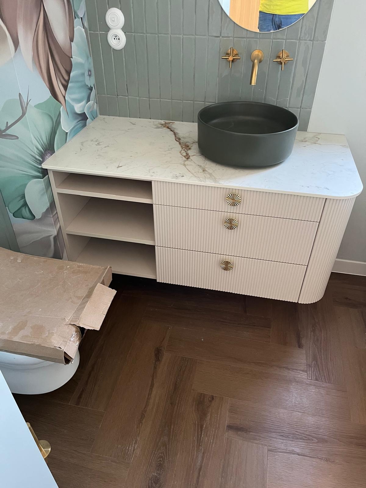 Łazienka z umywalką nablatową w kolorze grafitowym, armaturą w odcieniu złota i szafką z marmurowym blatem oraz ryflowanymi frontami szuflad, widoczny karton po ceramice i parkiet w jodełkę.