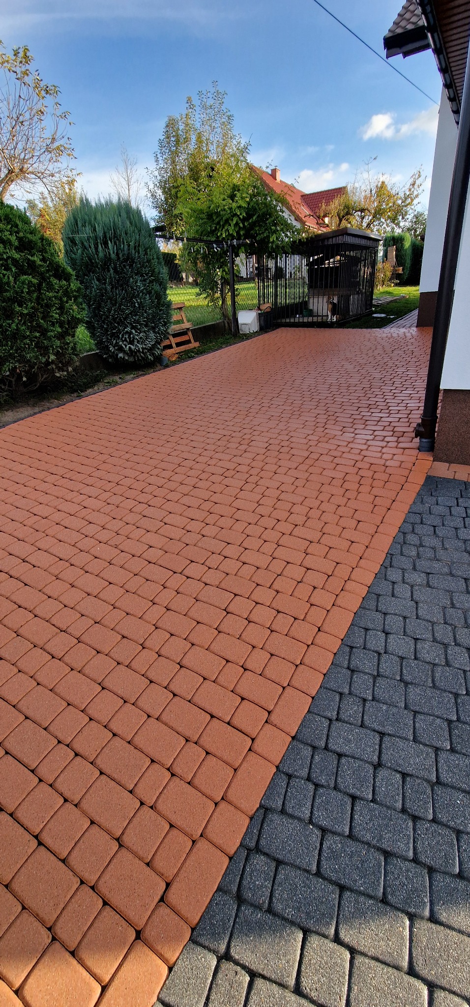 Ułożona kostka brukowa w kolorze terakoty kontrastująca z szarą kostką, widok na zadbany ogród z klatką dla psa w tle.