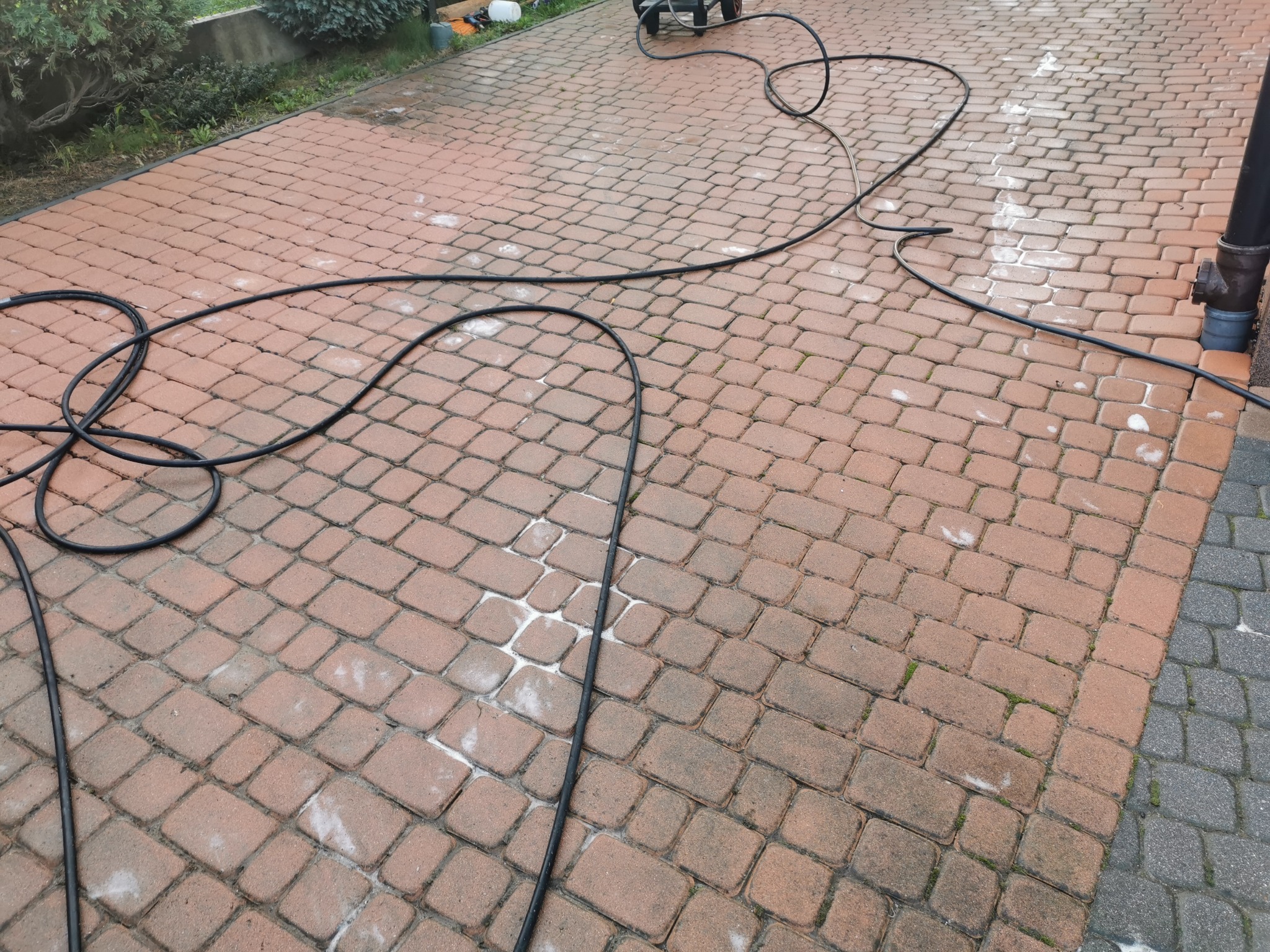 Czerwona kostka brukowa częściowo oczyszczona, widoczne ślady po myciu ciśnieniowym, czarny wąż ciśnieniowy leżący na powierzchni.