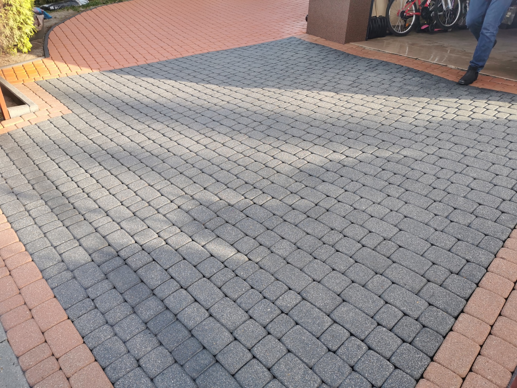 Ułożona szara kostka brukowa z obramowaniem z czerwonej kostki, widoczne fragmenty elewacji budynku i zaparkowane rowery