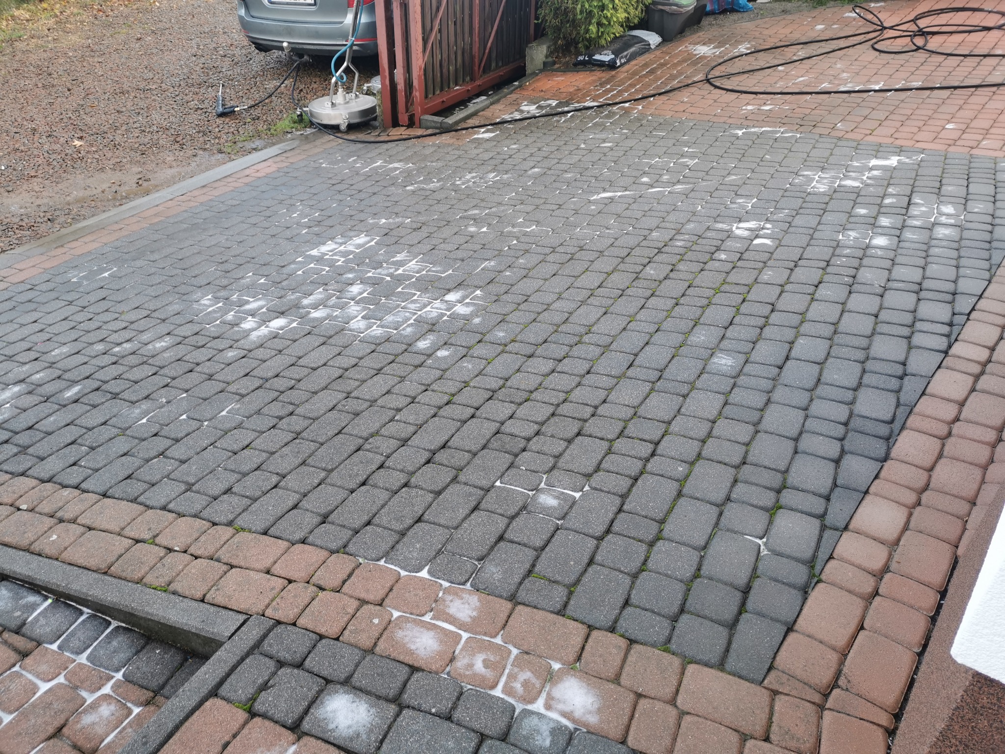 Fragment kostki brukowej w trakcie czyszczenia, widoczne ślady po umyciu oraz urządzenie do mycia pod ciśnieniem w tle.