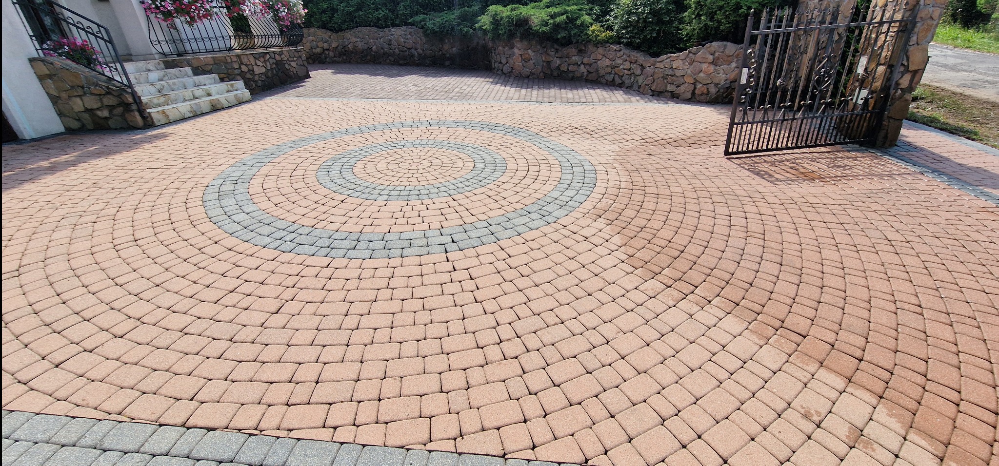 Ułożona z kostki brukowej, okrągła powierzchnia podjazdu w odcieniach brązu i szarości, z centralnym wzorem koła, prowadząca do metalowej bramy i kamiennego murku.
