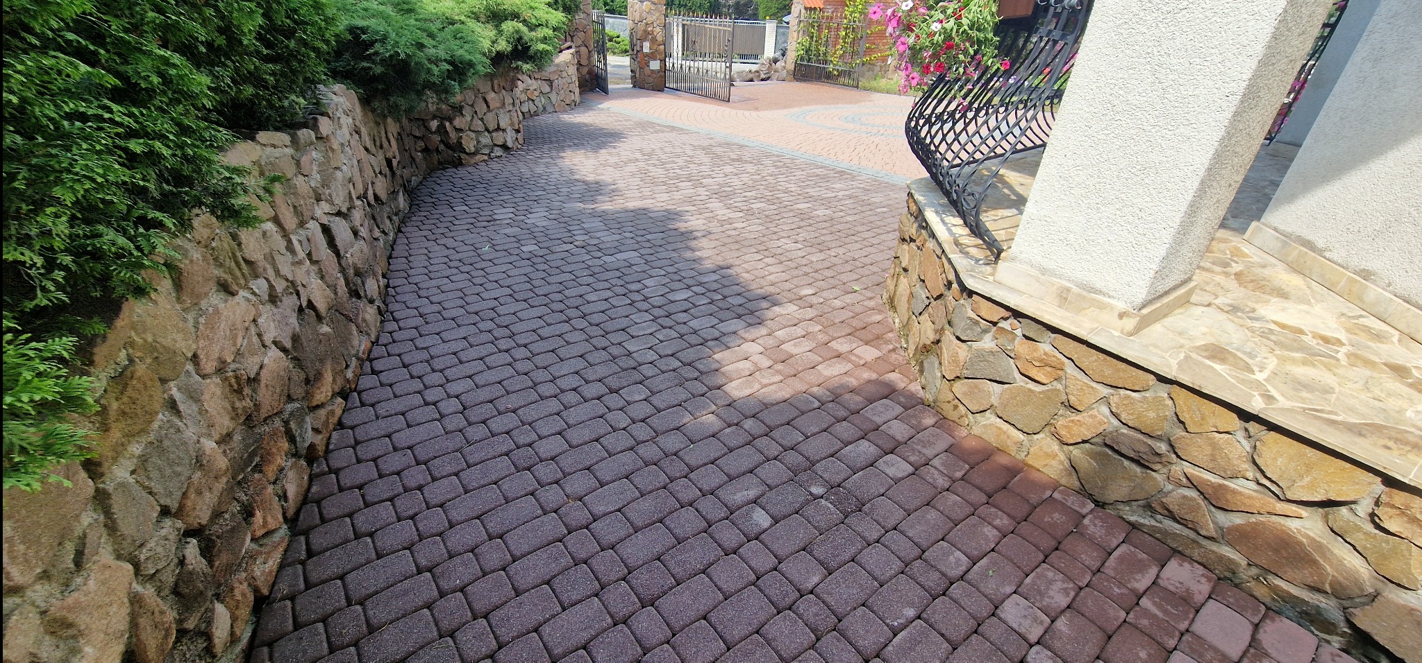 Ciemnobrązowa kostka brukowa ułożona na podjeździe, obramowana kamiennym murem i zielenią, prowadząca do metalowej bramy w tle.