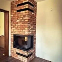 Narożny wkład opalany drewnem, obudowany czerwoną cegłą z czarnymi, poziomymi akcentami, ustawiony na drewnianej podłodze w rogu jasnego pomieszczenia.