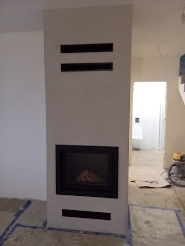 Nowoczesny, minimalistyczny wkład kominkowy z czarną ramą, w trakcie montażu w surowym wnętrzu. Widoczne dwie czarne poziome szczeliny wentylacyjne nad wkładem i jedna pod nim. W tle widoczna...