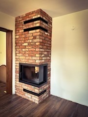 Narożny wkład opalany drewnem, obudowany czerwoną cegłą z czarnymi, poziomymi akcentami, ustawiony na drewnianej podłodze w rogu jasnego pomieszczenia.