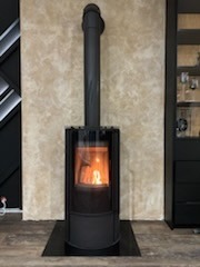 Wolnostojący piec opalany drewnem z przeszklonym paleniskiem, widoczny płomień, czarna obudowa i rura odprowadzająca dym, na tle beżowej ściany z dekoracyjną fakturą.