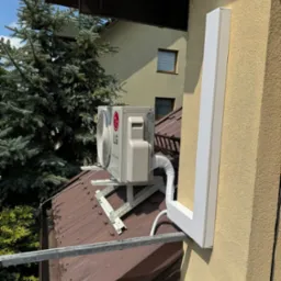 Zewnętrzna jednostka klimatyzacji LG Dual Inverter zamontowana na wsporniku na dachu domu, z widoczną osłoną instalacji na elewacji budynku.
