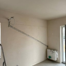 SV CLIMA Serhii Vyhorchuk - Wnętrze w trakcie prac instalacyjnych: rury poprowadzone po ścianie, nowy grzejnik zabezpieczony folią, widoczna drabina malarska i ślady po kuciu w ścianie.