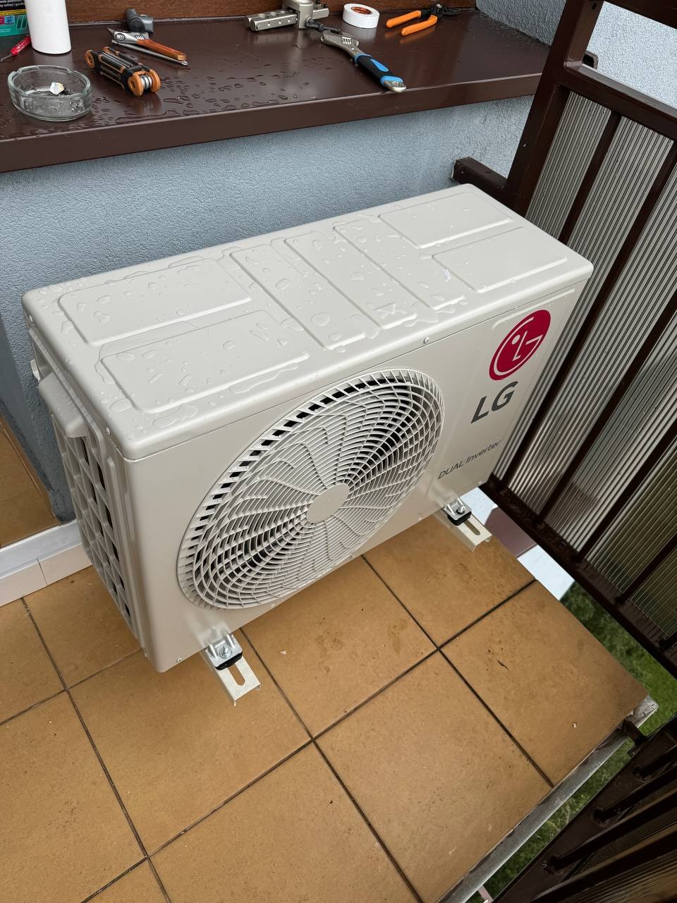 Zewnętrzna jednostka klimatyzacji LG Dual Inverter zamontowana na brązowych płytkach tarasowych, widoczne ślady wilgoci i narzędzia na parapecie w tle.