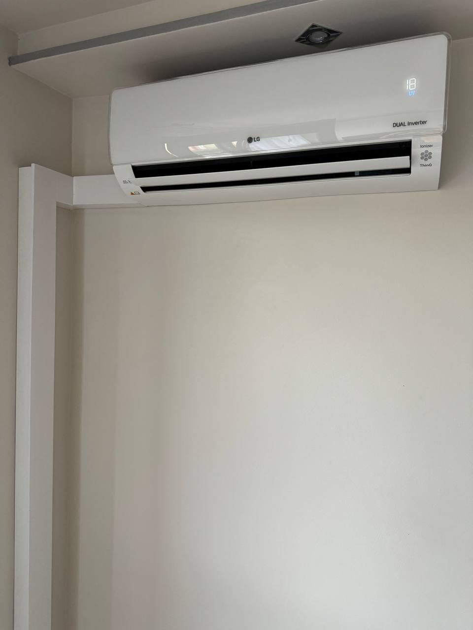 Biały klimatyzator LG Dual Inverter z wyświetlaczem ustawionym na 18 stopni, zamontowany na ścianie w jasnym pomieszczeniu, widoczny element instalacji elektrycznej w suficie.