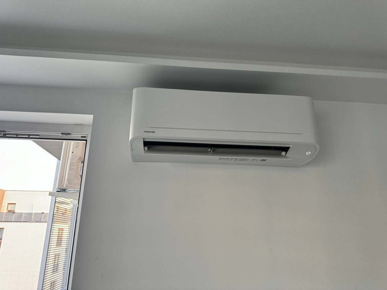 Biała klimatyzacja Toshiba zamontowana na jasnej ścianie w minimalistycznym wnętrzu, widoczna część okna z żaluzjami w tle.