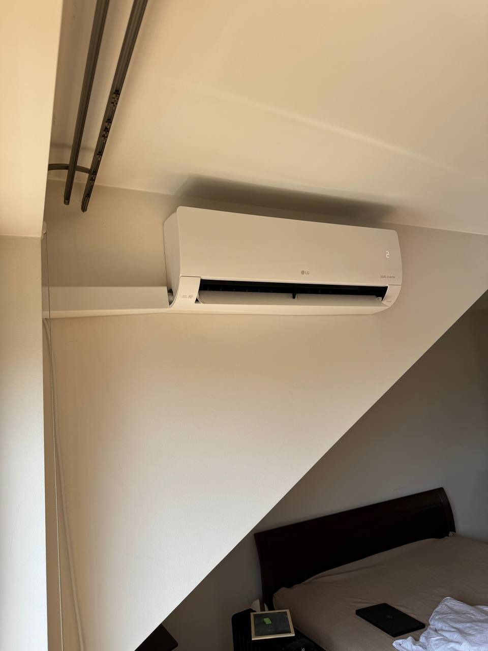 Biały klimatyzator LG zamontowany na ścianie w pokoju na poddaszu, widoczne metalowe rury pod sufitem i fragment łóżka.