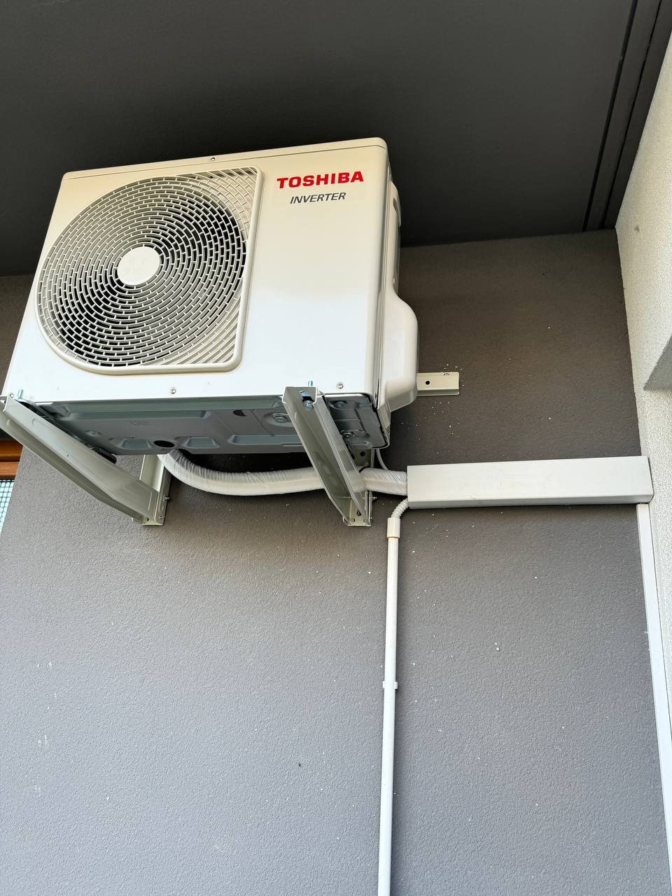 Zewnętrzna jednostka klimatyzacji Toshiba Inverter zamontowana na szarej ścianie budynku, widoczne białe rury odprowadzające skropliny i maskownica przewodów.